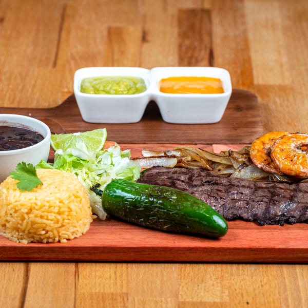 Grilled steak and shrimp with rice, black beans, salad, jalapeño, and sauces on a wooden board.