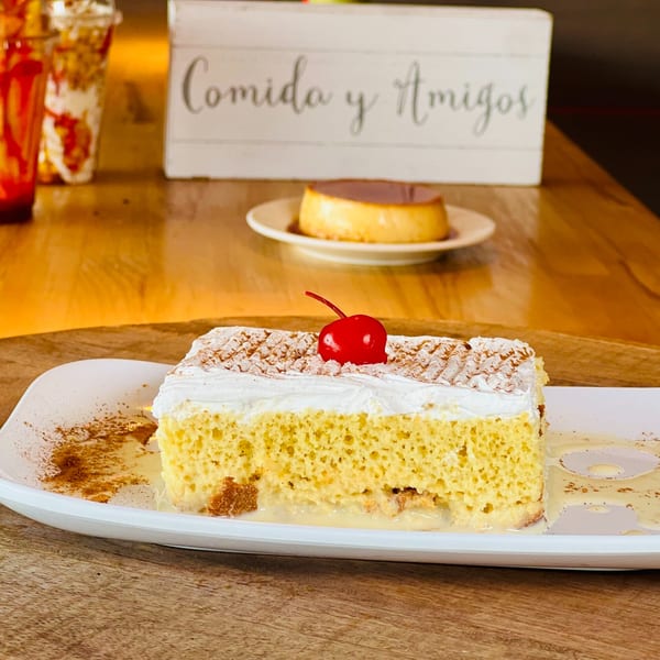 Slice of tres leches cake with whipped cream and a cherry, with flan in the background.