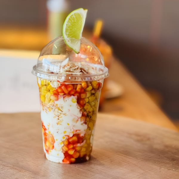 Elote loco in a clear plastic cup, topped with a lime wedge, on a wooden table.