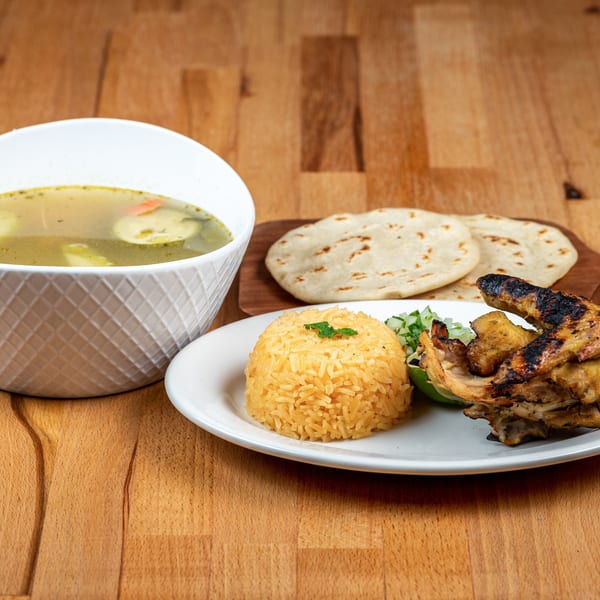 Salvadoran meal with chicken, yellow rice, tortillas, and a bowl of vegetable soup on a wooden table.