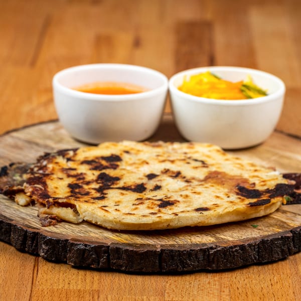 A charred pupusa on a wooden board, with two small white bowls of curtido and salsa.